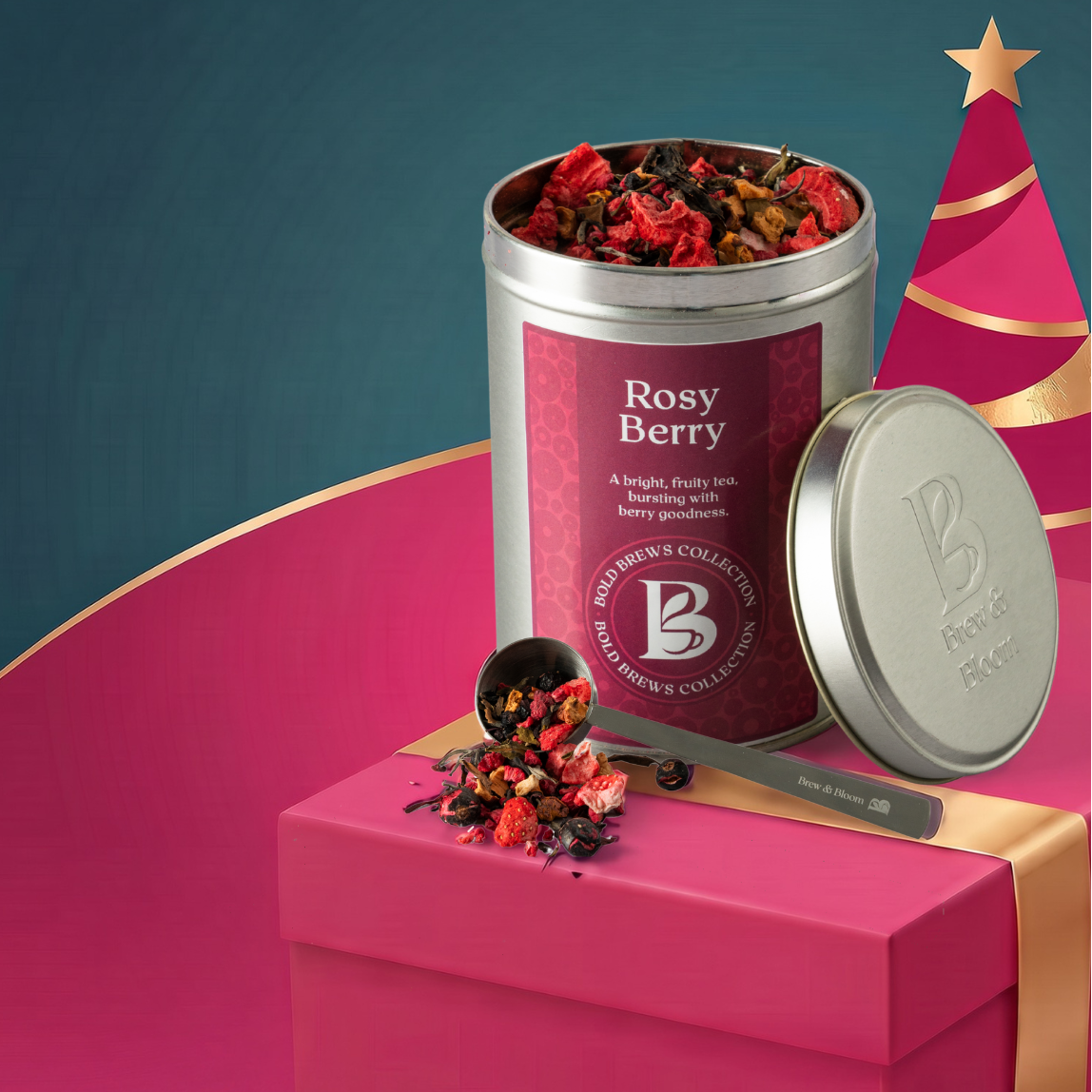 A tin of Rosy Berry fruit tea from Brew & Bloom is displayed next to an open lid on a bright pink gift box with gold ribbon, set against a teal background with a pink Christmas tree graphic.