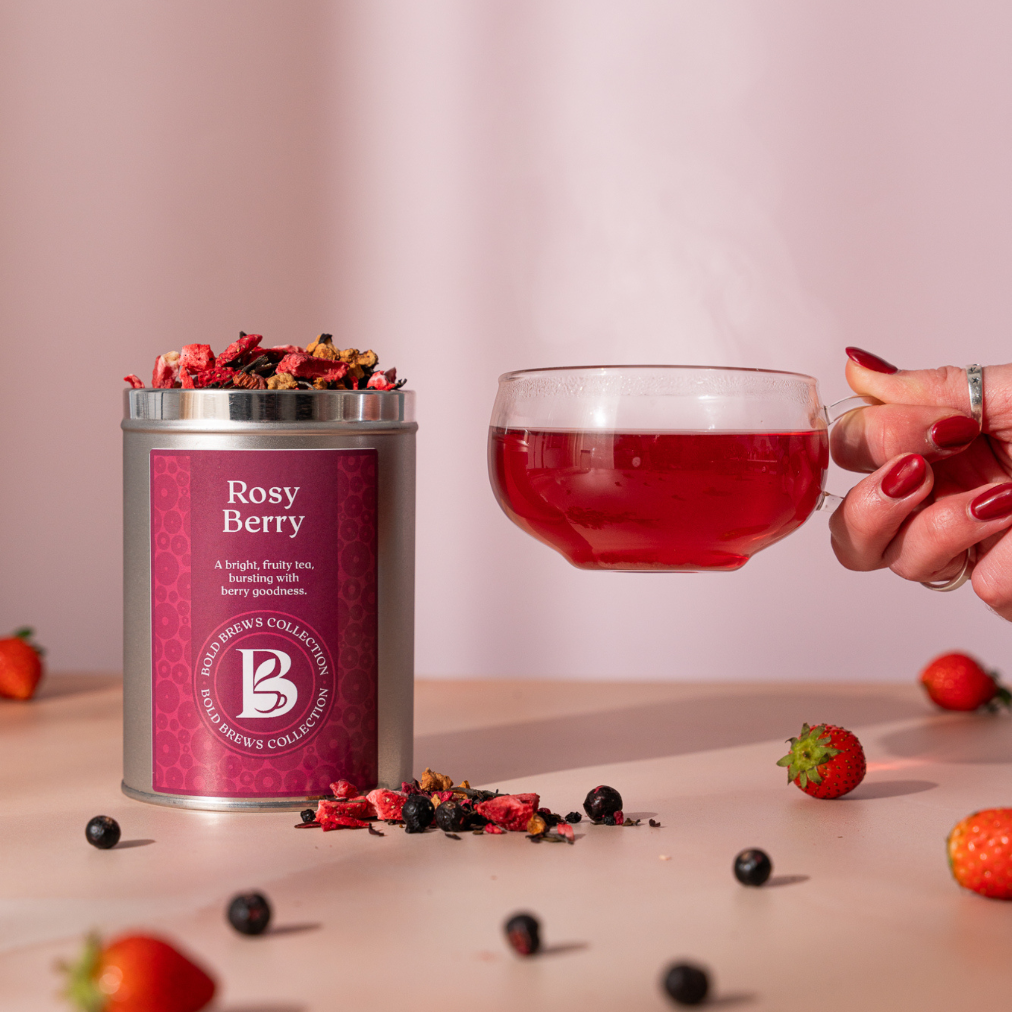 Cup of Rosy Berry fruit tea beside Brew & Bloom tin with fresh berries on soft pink background